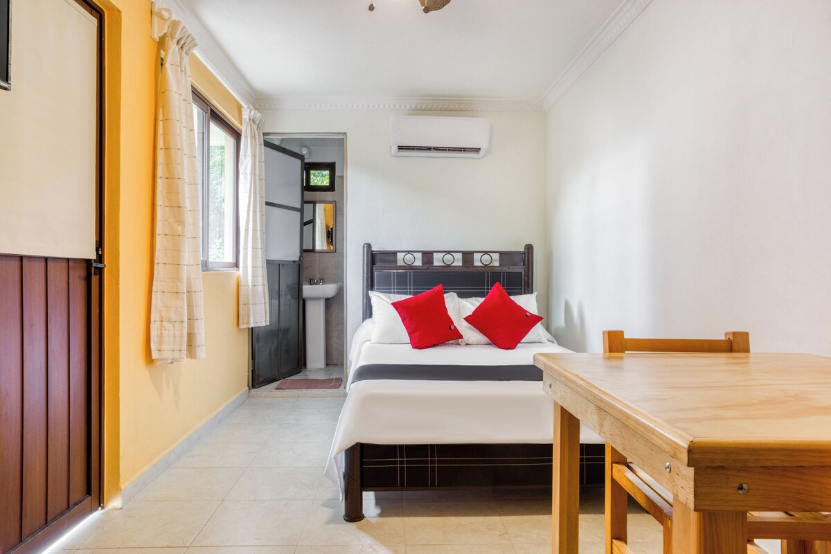 A bedroom with a bed, a wooden table, a bathroom with a sink, and an air conditioning unit.