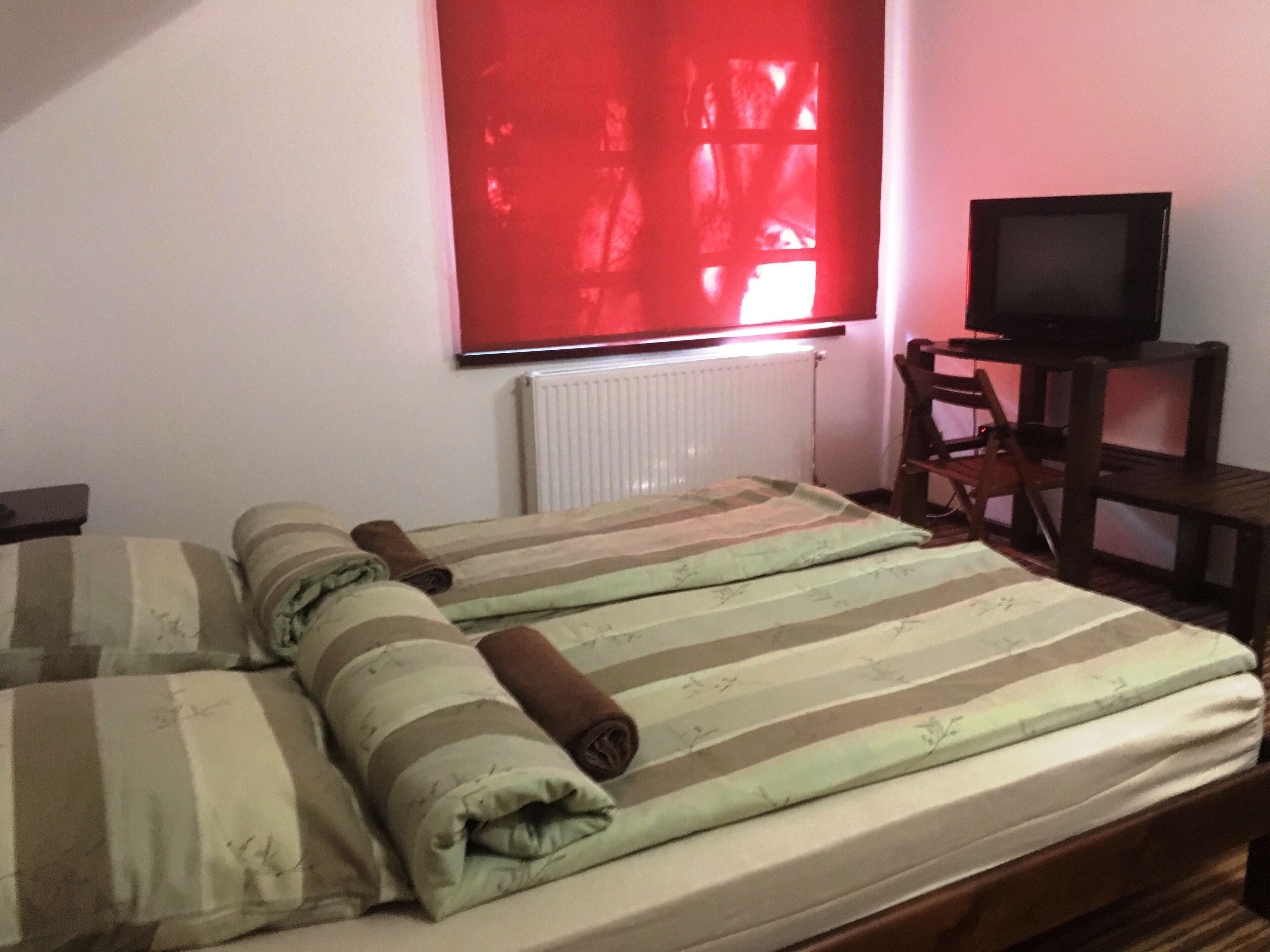 Un lit simple avec une literie à rayures et des serviettes enroulées, un téléviseur posé sur un meuble en bois, et un store rouge à la fenêtre.