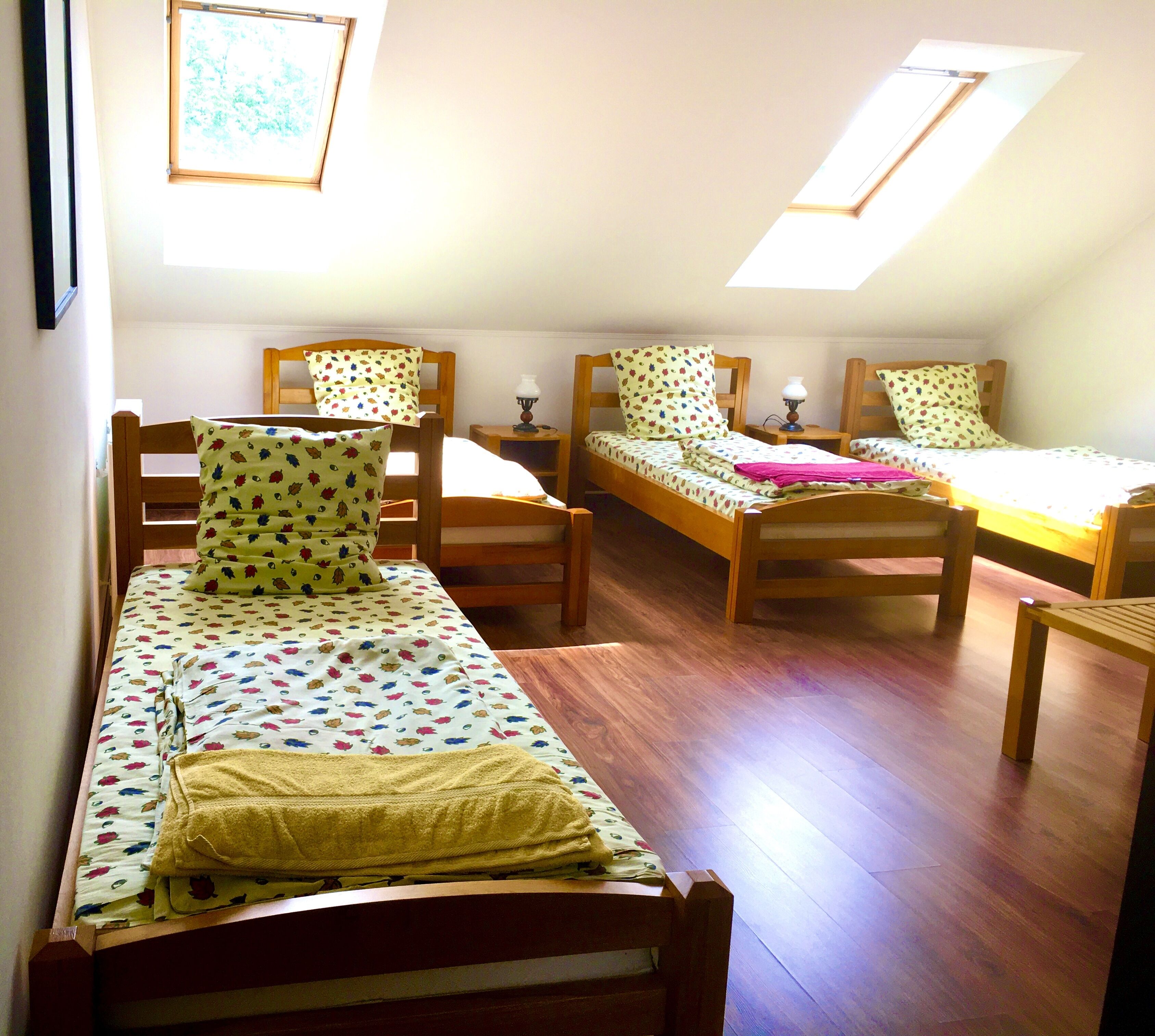 A dormitory room with three wooden bunk beds, each with a matching headboard and footboard. There are two bedside tables with lamps, and the room has a sloped ceiling with skylights.
