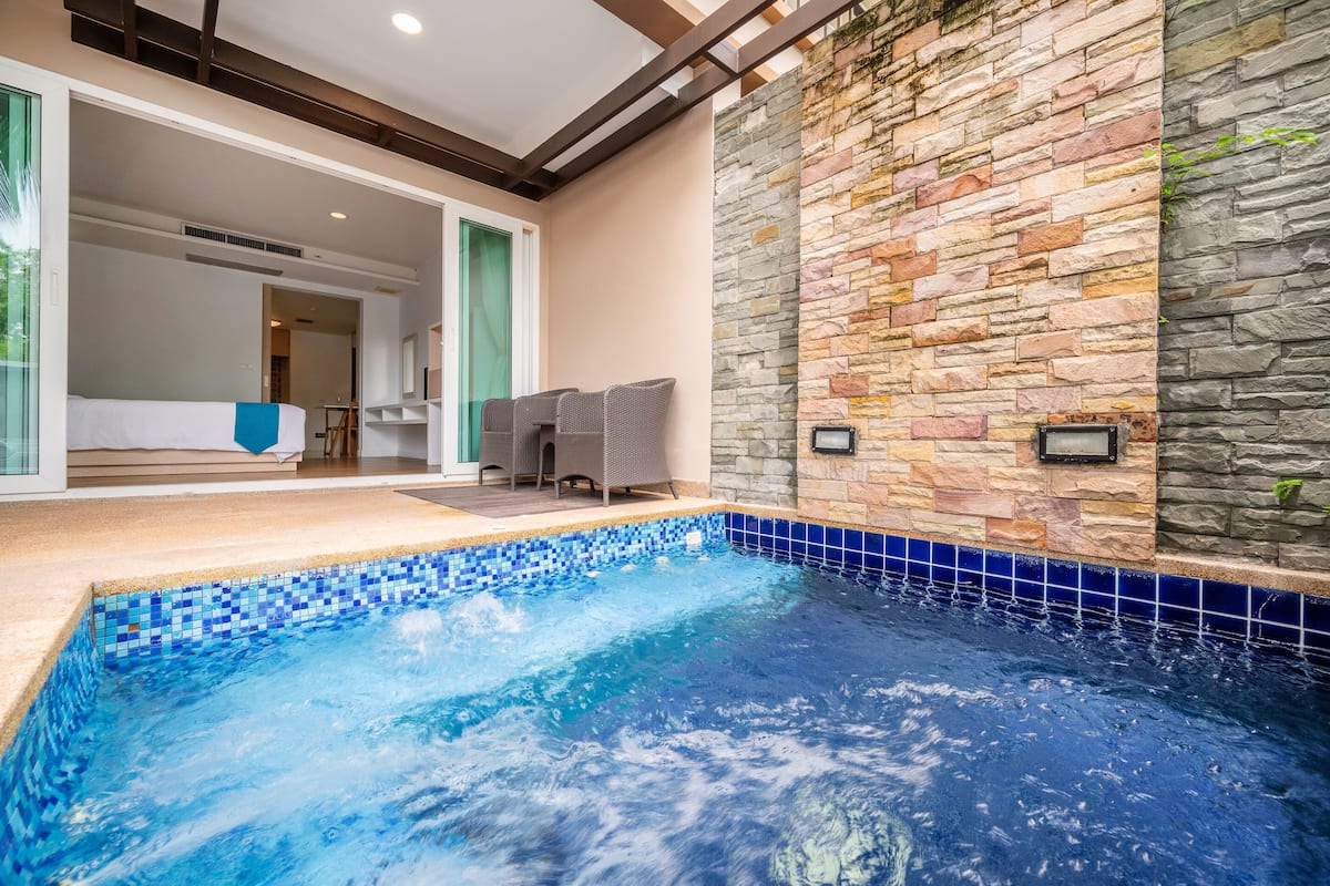Una piscina interior moderna con bordes de mosaicos, paredes de piedra y una zona de estar con sillas de mimbre.