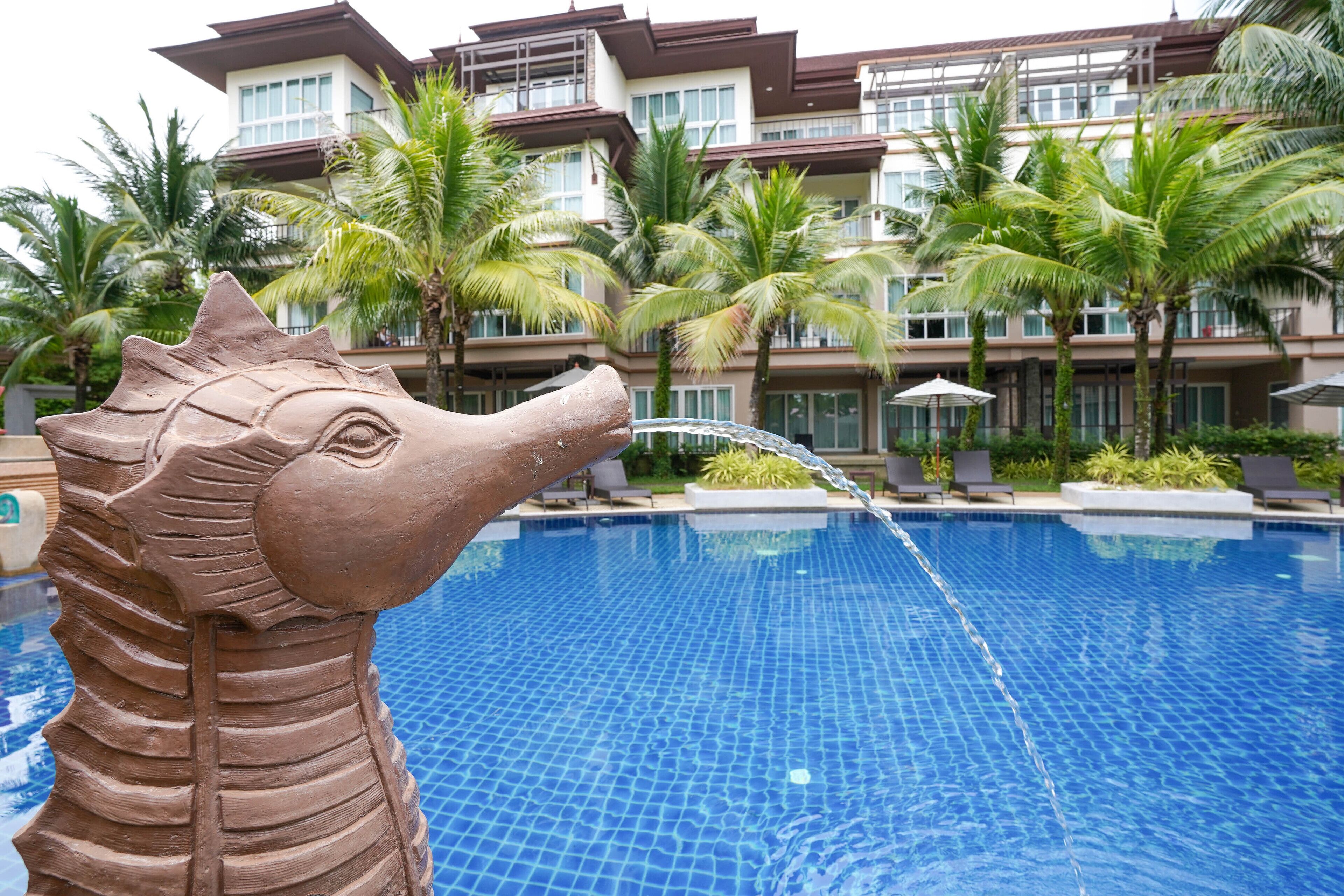 A swimming pool with a seahorse sculpture, palm trees, and a multi-story building in the background.