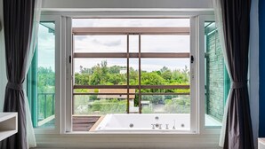 Un dormitorio con un amplio ventanal corredera que da a un balcón, una bañera y vistas a zonas verdes y edificios.