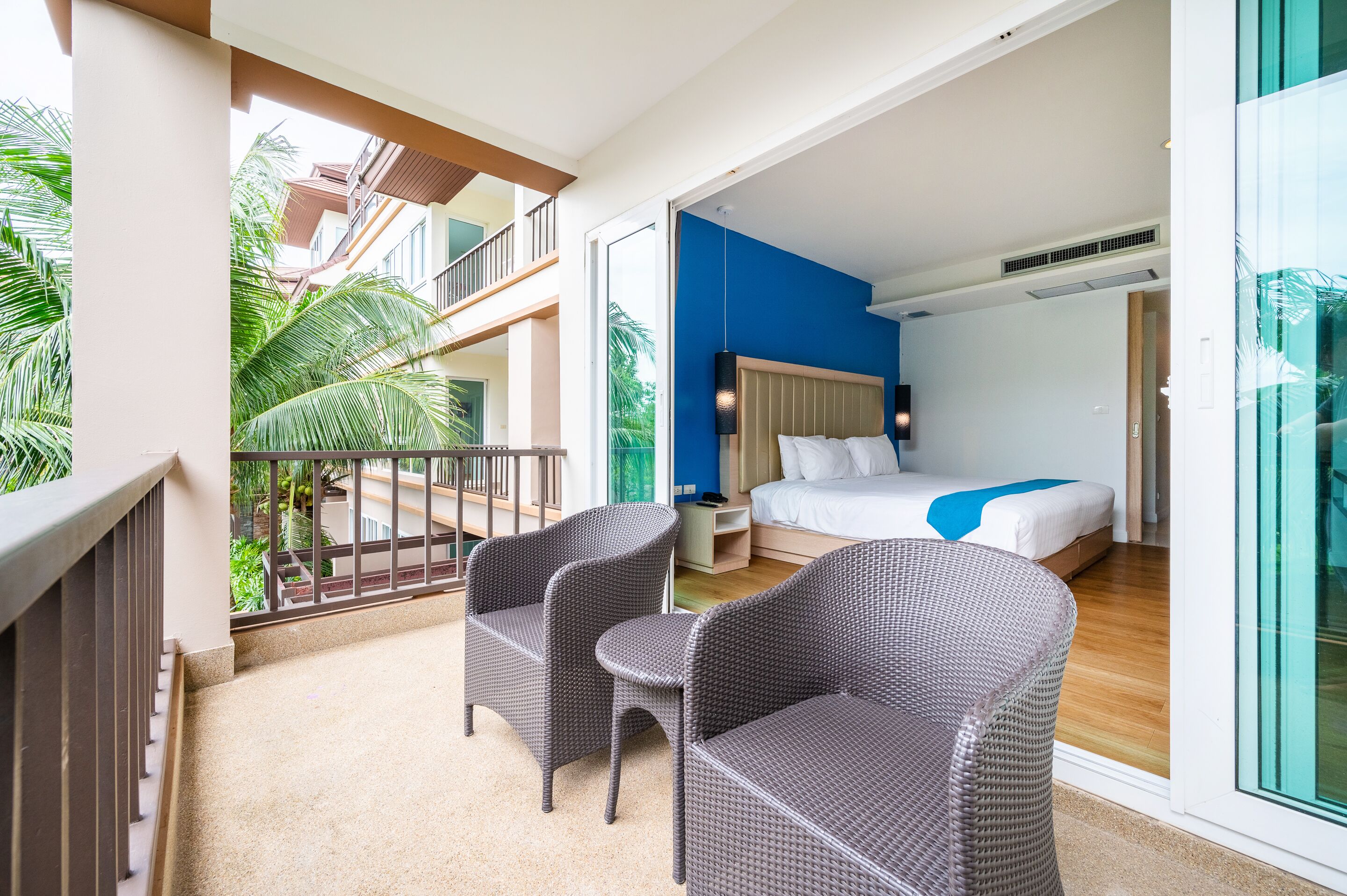 Un balcón con mobiliario de mimbre, un dormitorio con cama y mesitas de noche, y vistas a un edificio y palmeras.
