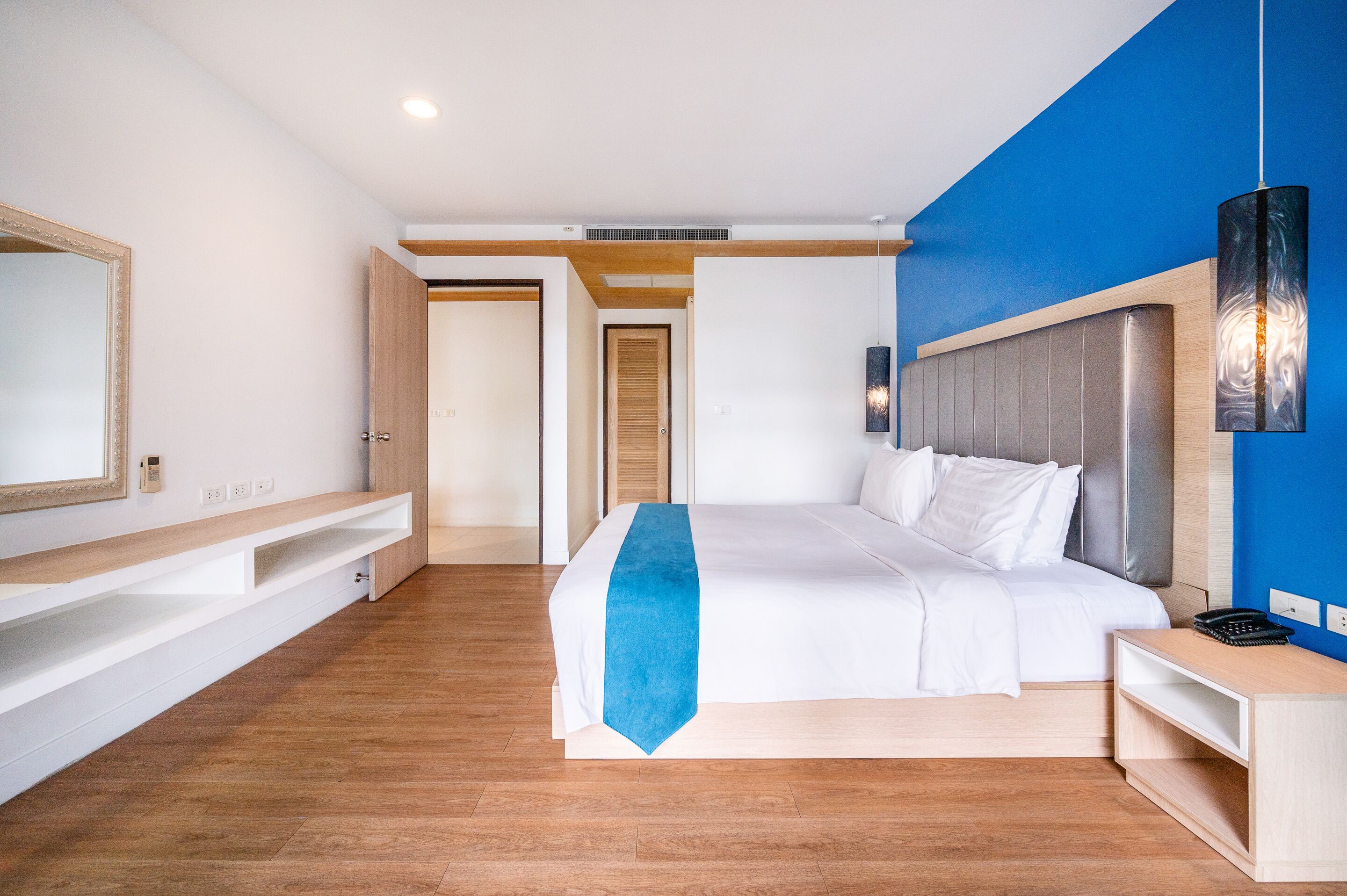 A modern hotel room with a large bed, a blue accent wall, wooden flooring, and built-in shelving.