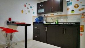 Una cocina compacta con armarios oscuros, un microondas y una pequeña encimera. Se observan adhesivos decorativos de café coloridos en la pared y una silla roja.