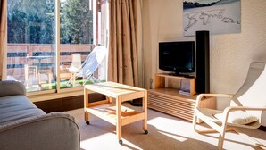 A living room with a sofa, a wooden coffee table, a TV on a stand, a chair, and a large window with curtains. - 3-room apartment near the slopes - Courchevel 1850 (Courchevel 1850)