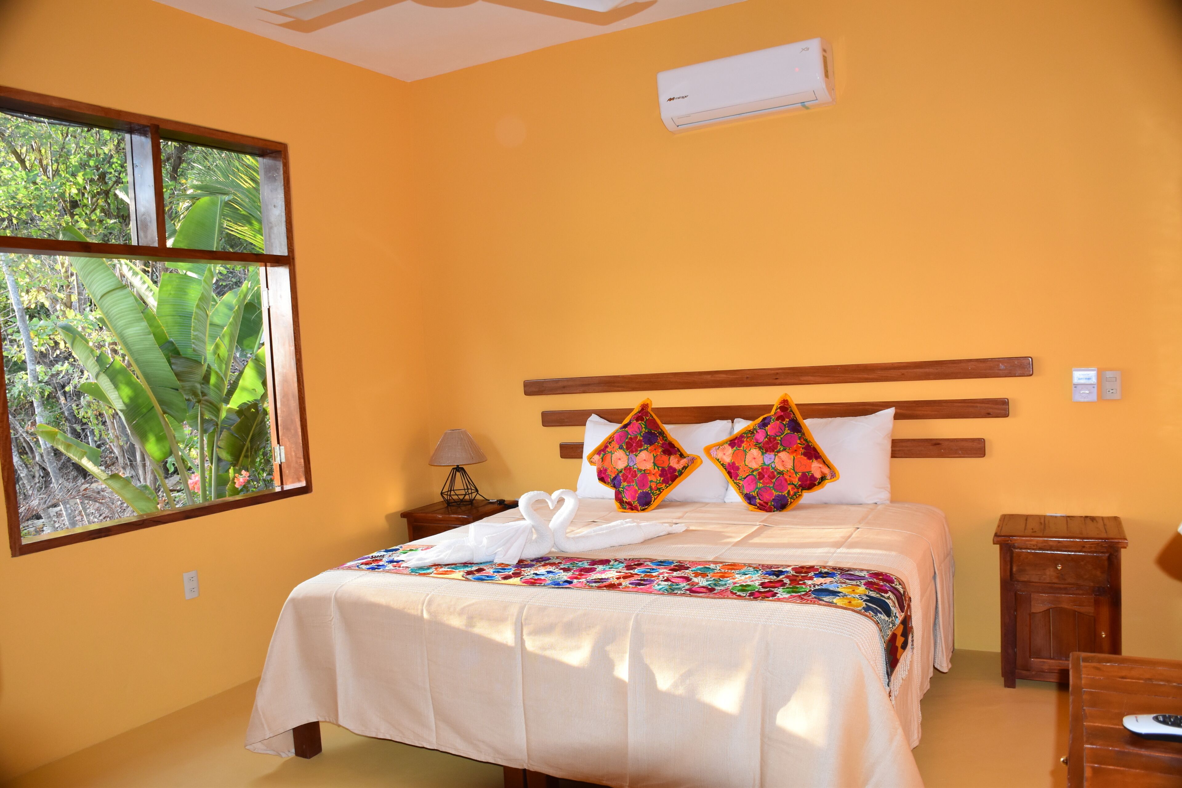 Una habitación de hotel con una cama, un ventanal con vistas a zonas verdes, una mesita de noche con una lámpara y un equipo de aire acondicionado en la pared.