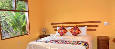 Una habitación de hotel con una cama, un ventanal con vistas a zonas verdes, una mesita de noche con una lámpara y un equipo de aire acondicionado en la pared.