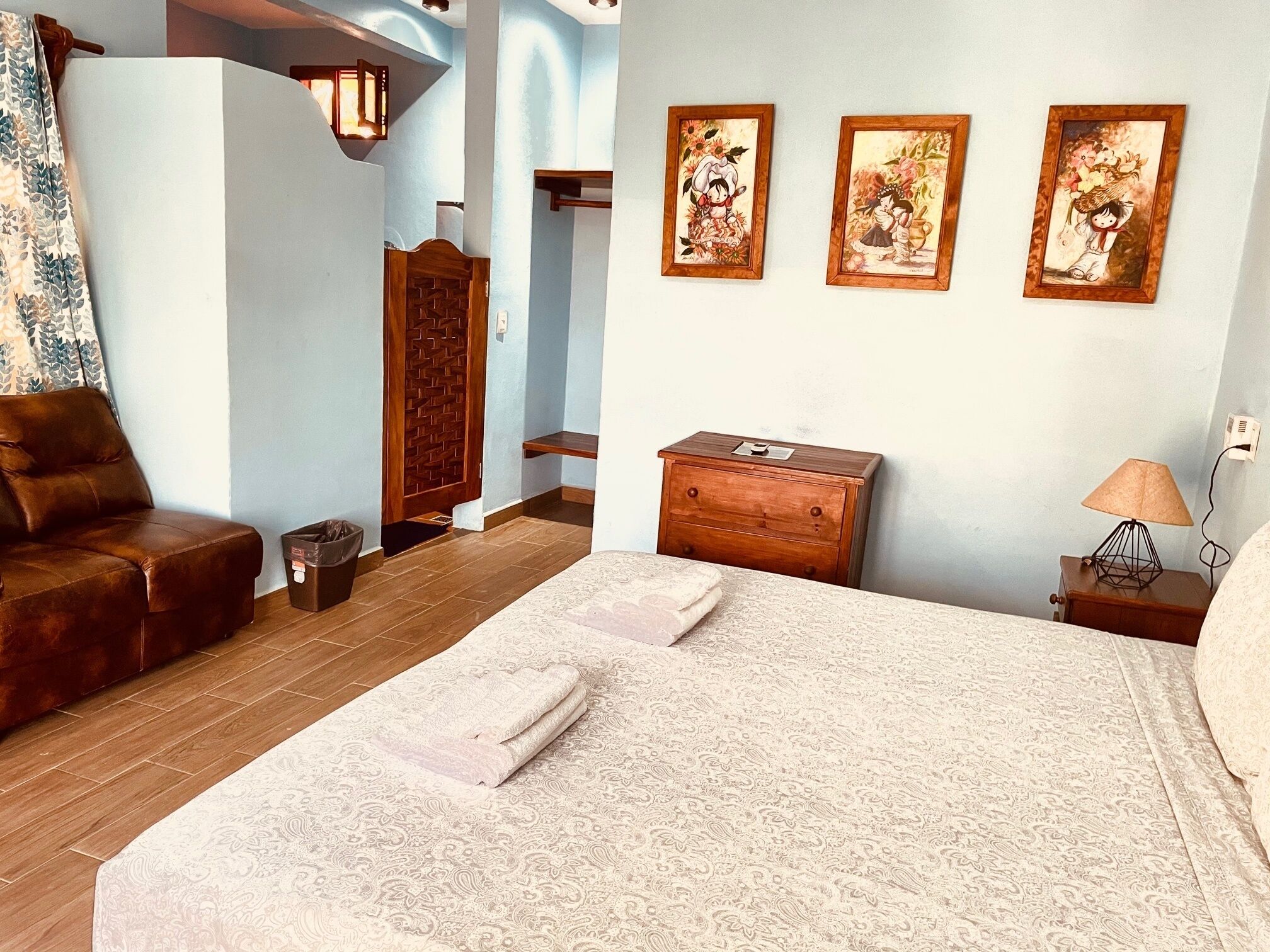 A bedroom with a bed, a wooden dresser, a nightstand with a lamp, and three framed pictures on the wall.