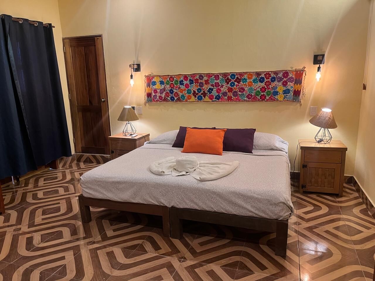 A neatly made bed with a patterned bedspread, two bedside lamps, a colorful tapestry on the wall, and a patterned floor.