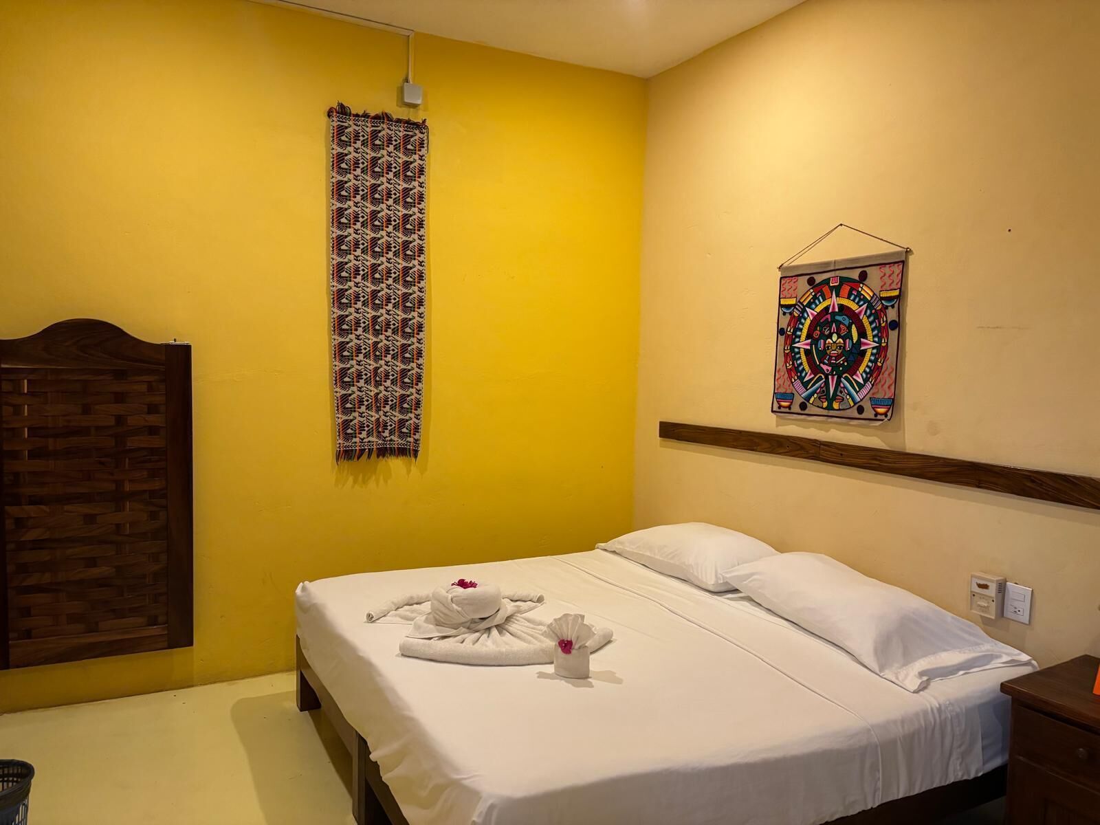 A hotel room with a bed, a wooden headboard, a yellow wall, a patterned curtain, and a colorful artwork.