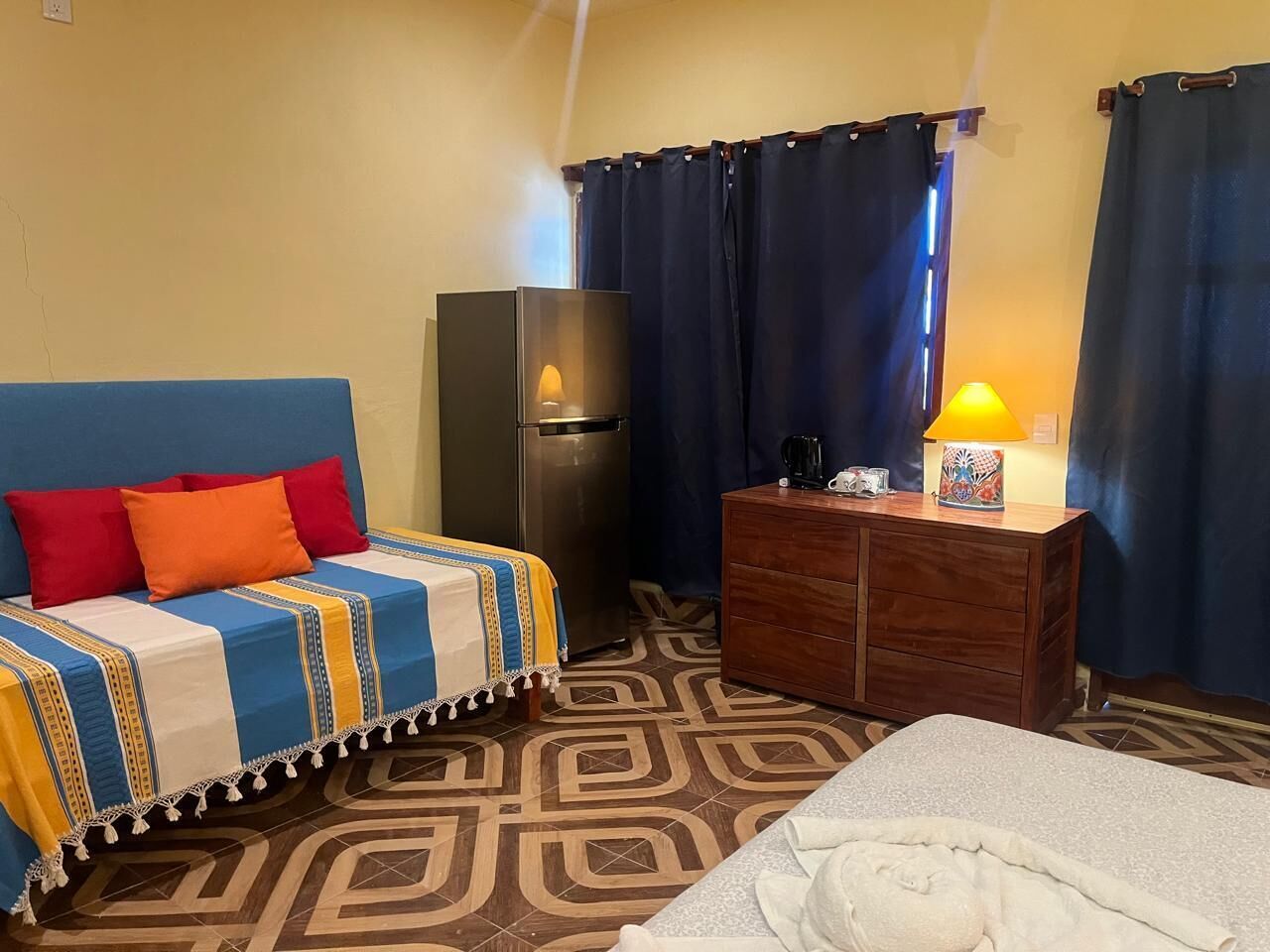 A hotel room with a bed, a sofa with colorful cushions, a wooden dresser, a refrigerator, and a patterned floor.