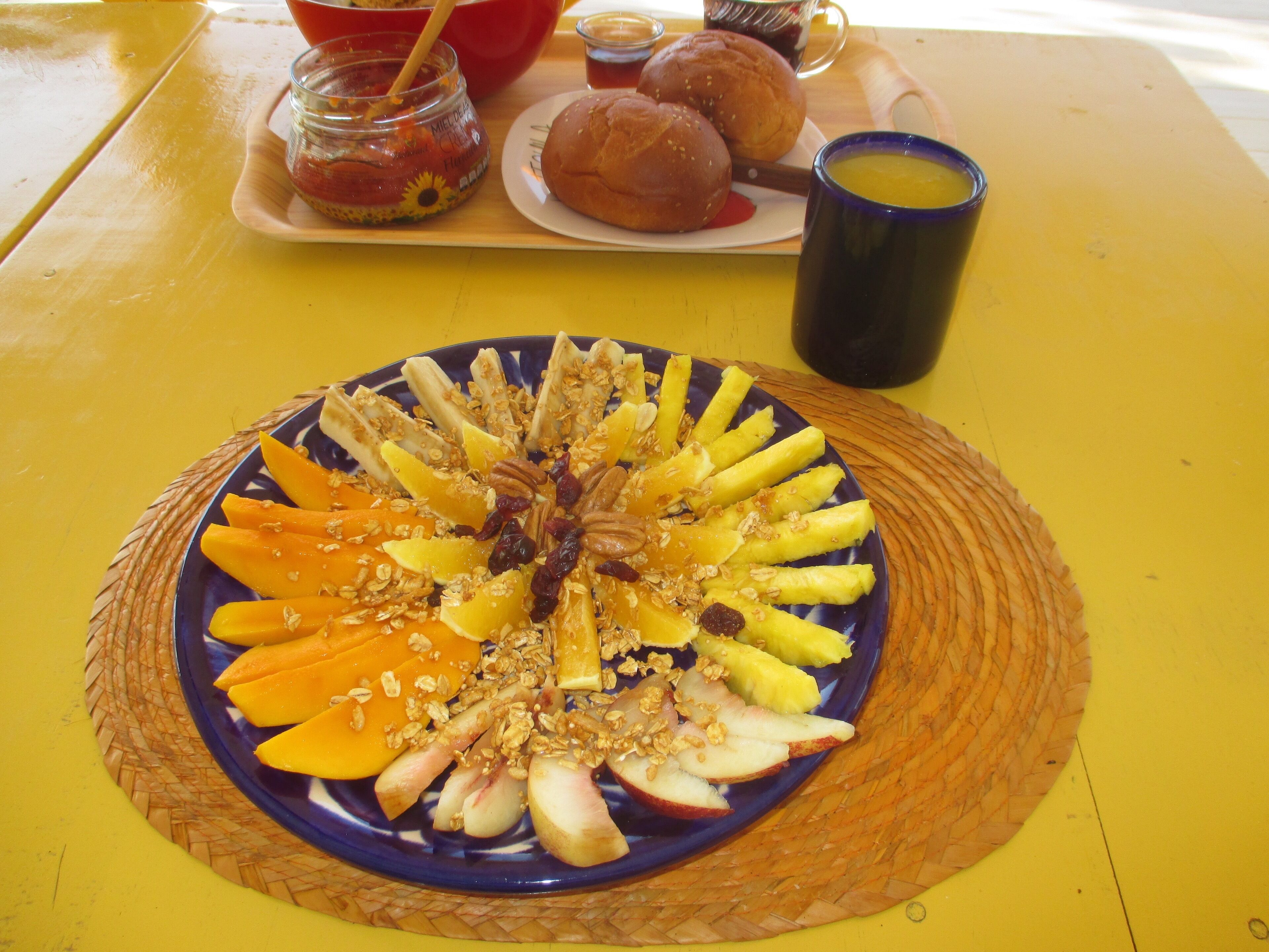 Un plato de desayuno con fruta y granola, una taza y un frasco de mermelada sobre una mesa amarilla.