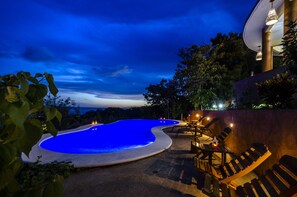 A swimming pool with a blue-lit edge, surrounded by lounge chairs and a dining area, set against a backdrop of trees and a clear sky. - Casa Biulù (San Pedro Pochutla)