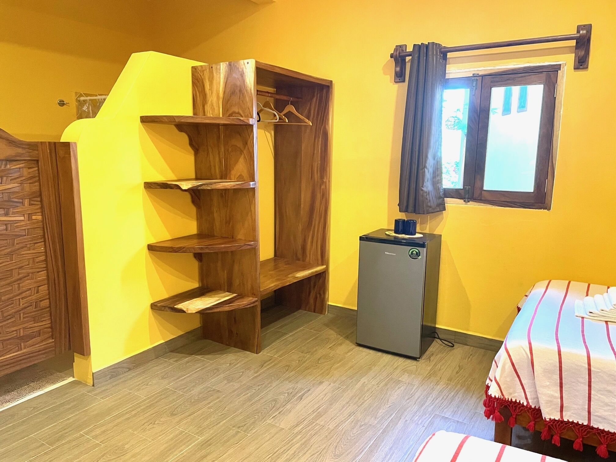 A room with a wooden wardrobe, a small refrigerator, a bed with a striped blanket, and a window with curtains.