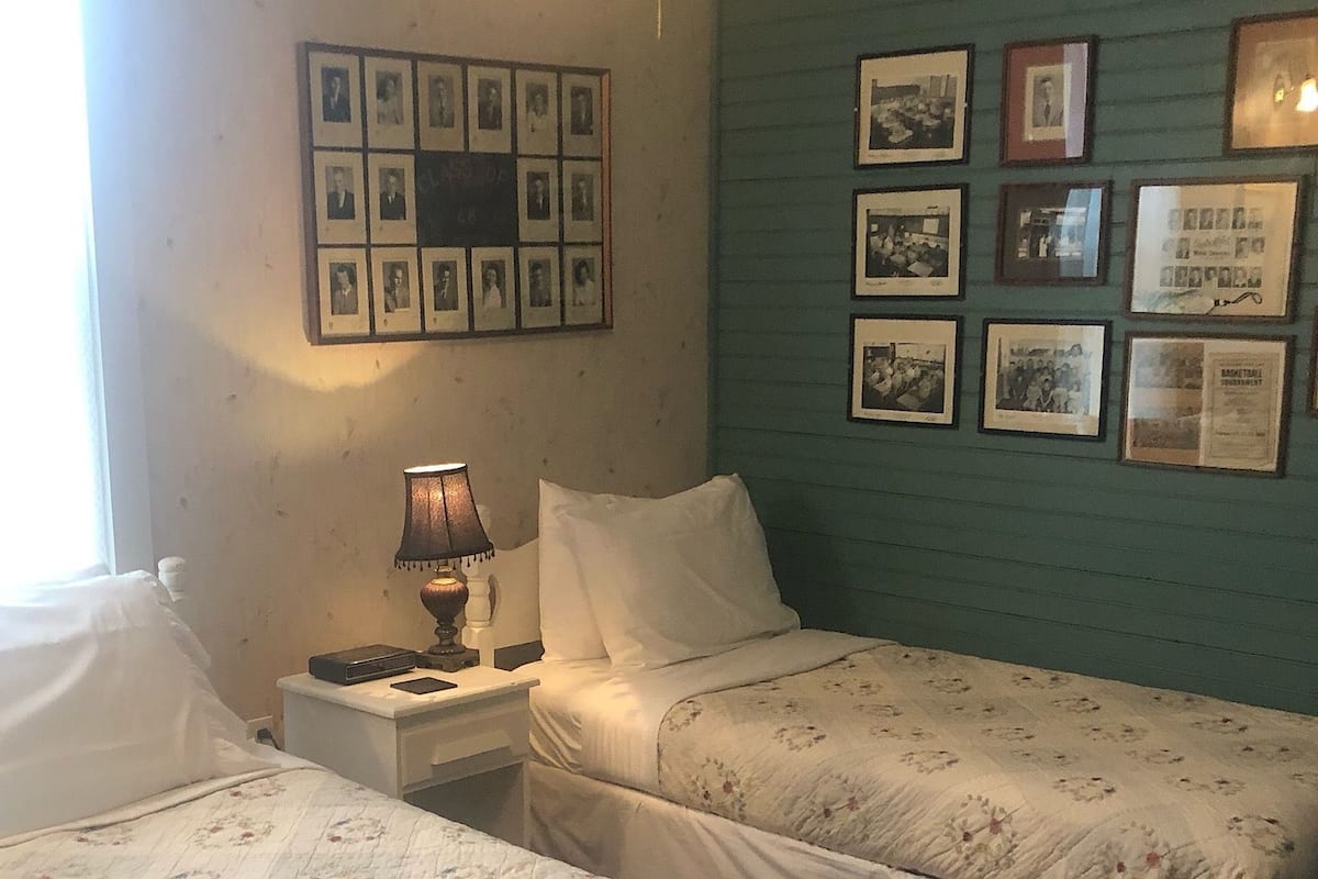 A room with two beds, a nightstand with a lamp, and a wall with framed pictures.