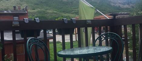 A balcony with two green metal chairs and a small round table, overlooking a parking area and a mountainous landscape.
