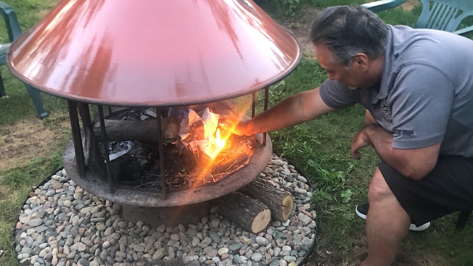 A man tending to a fire in a metal fire pit with a conical roof, surrounded by pebbles.