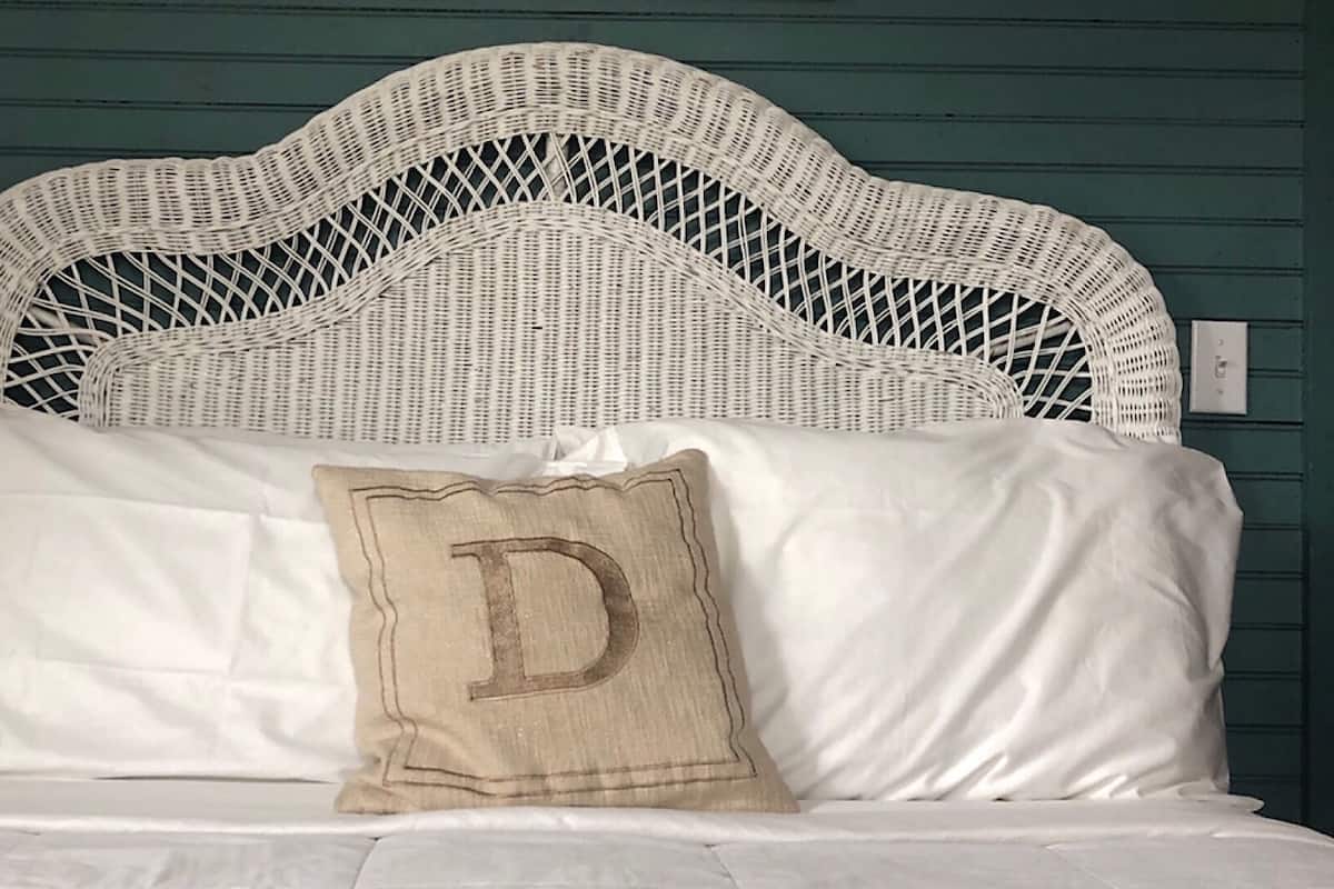A bed with a white wicker headboard, white bedding, and a pillow with a large 'D' on it.