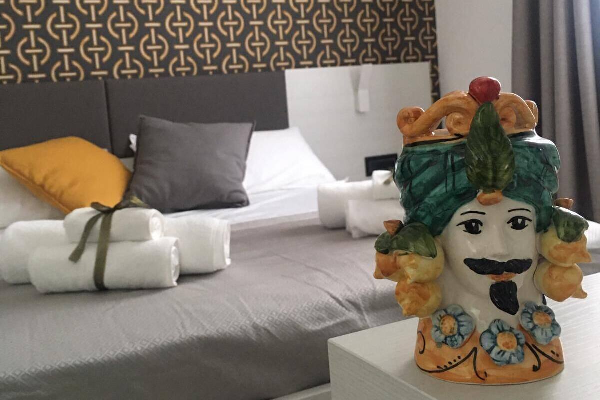 Une chambre d’hôtel avec un papier peint à motifs, un lit avec du linge de maison blanc, un oreiller gris et une figurine décorative en céramique posée sur une table de chevet.