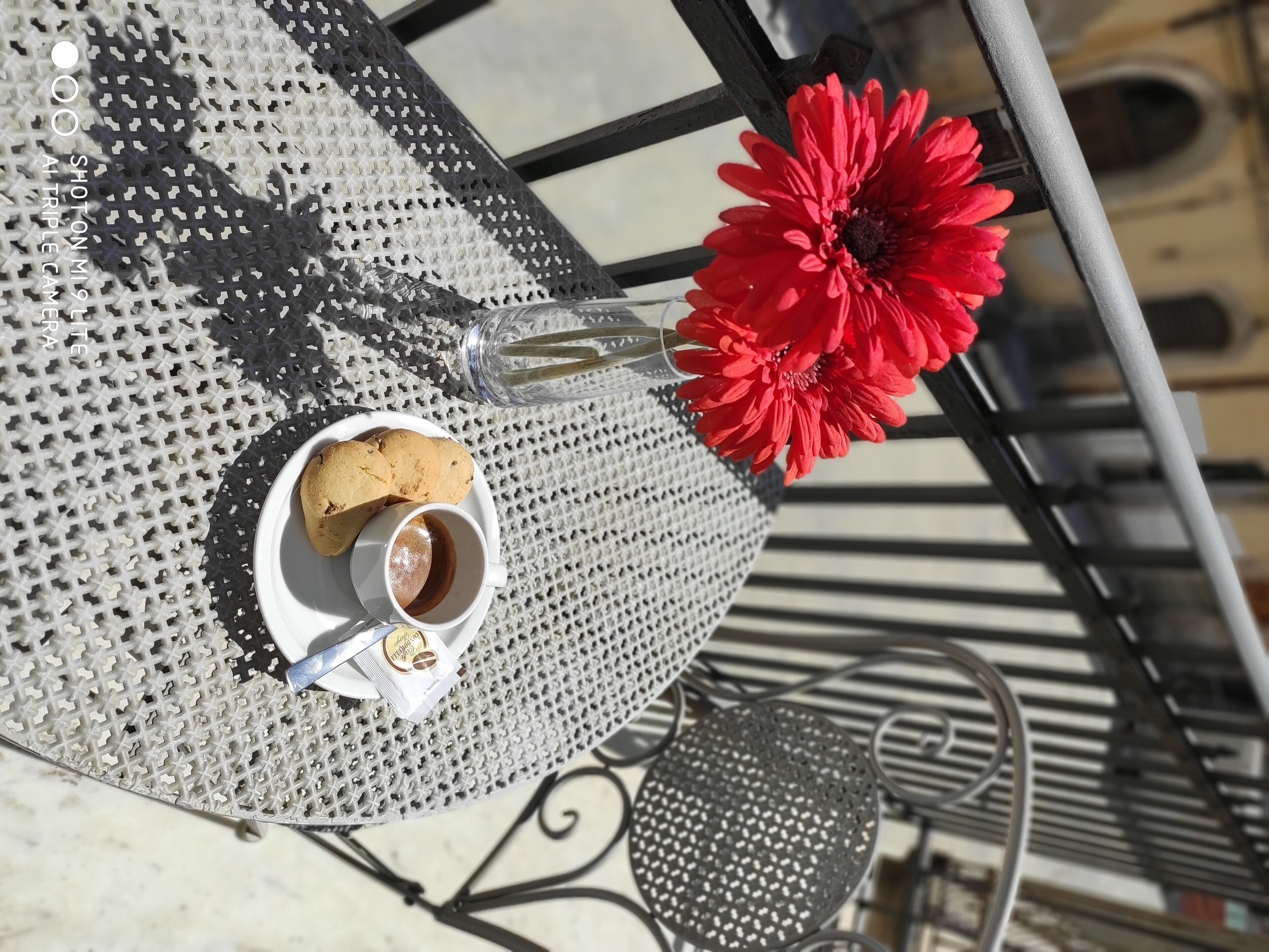 Une table sur laquelle se trouve une assiette blanche contenant une pâtisserie et une tasse de café, un verre transparent et une fleur rouge.