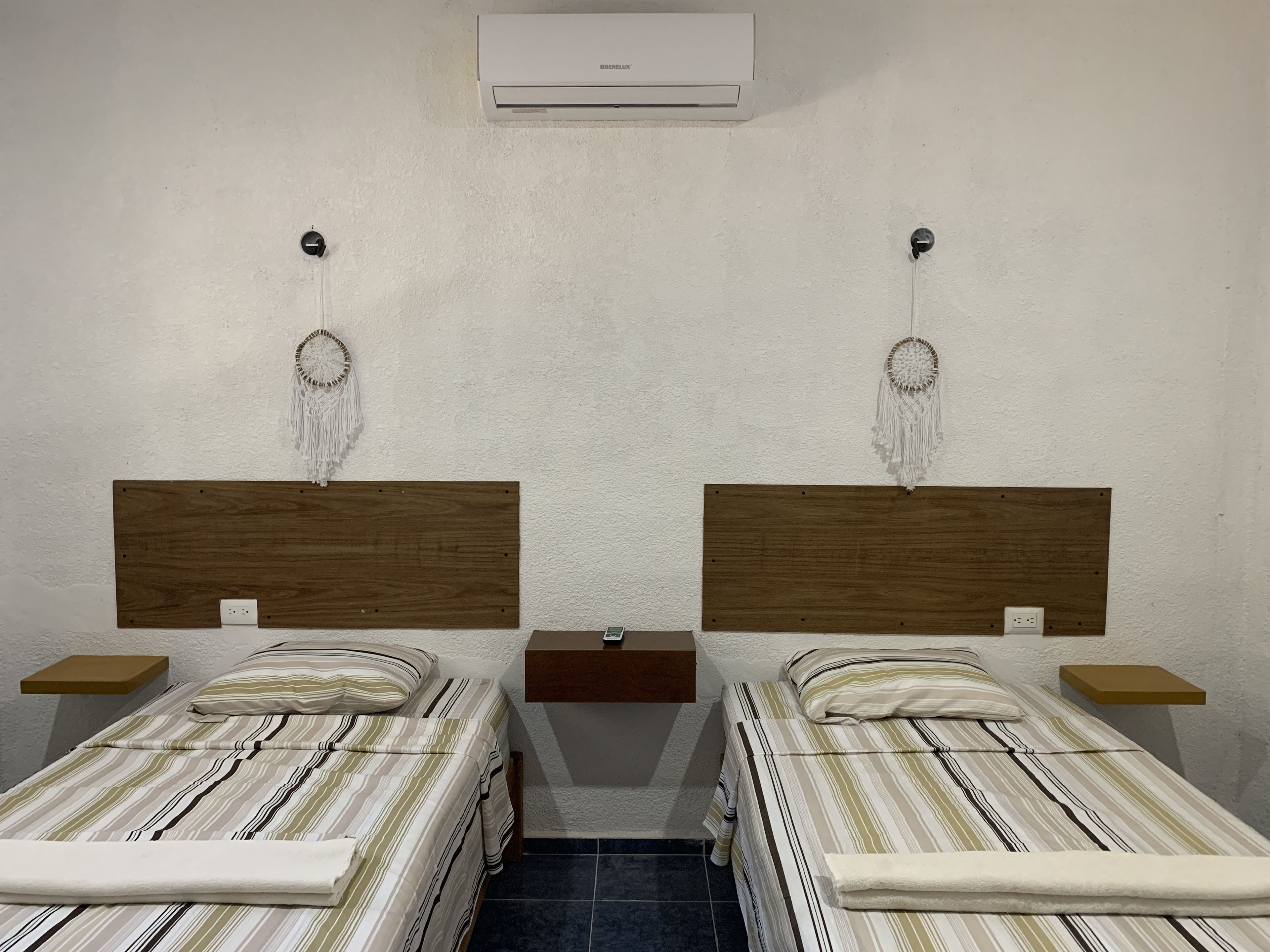 A room with two single beds, each with a wooden headboard and a bedside table. There is a wall-mounted air conditioner above the beds.
