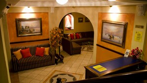 A lobby with a reception desk, two sofas with red and beige cushions, a large vase of flowers, and two framed paintings on the walls. - Hotel Merliot (Antiguo Cuscatlán)
