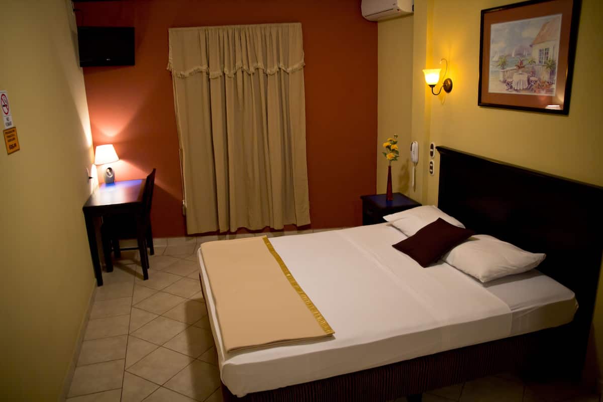 Ein Hotelzimmer mit einem Bett, einem Schreibtisch mit Lampe, einem Stuhl, einem Nachttisch, einem Gemälde und einem Fenster mit Vorhängen.