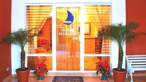 A hotel entrance with a large wooden door, a crescent moon logo, and two potted plants on either side. - Hotel Merliot (Antiguo Cuscatlán)