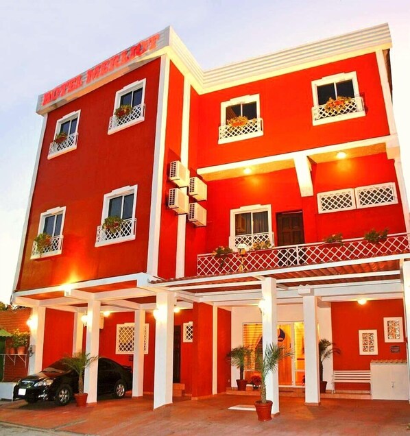 A red hotel with multiple balconies and air conditioning units. - Hotel Merliot (Antiguo Cuscatlán)