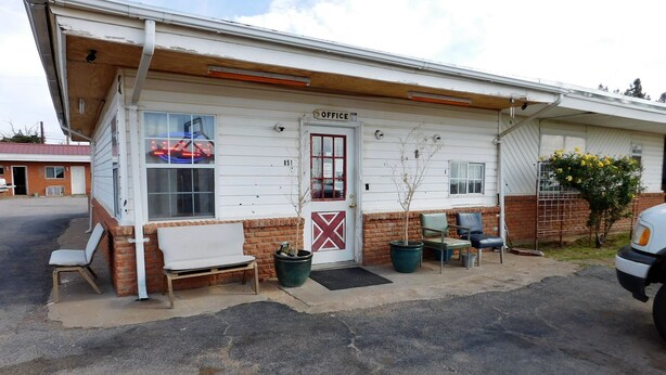 A small building with a sign reading "OFFICE" and a door marked with a red cross. There are two chairs outside, a potted plant, and a small tree.