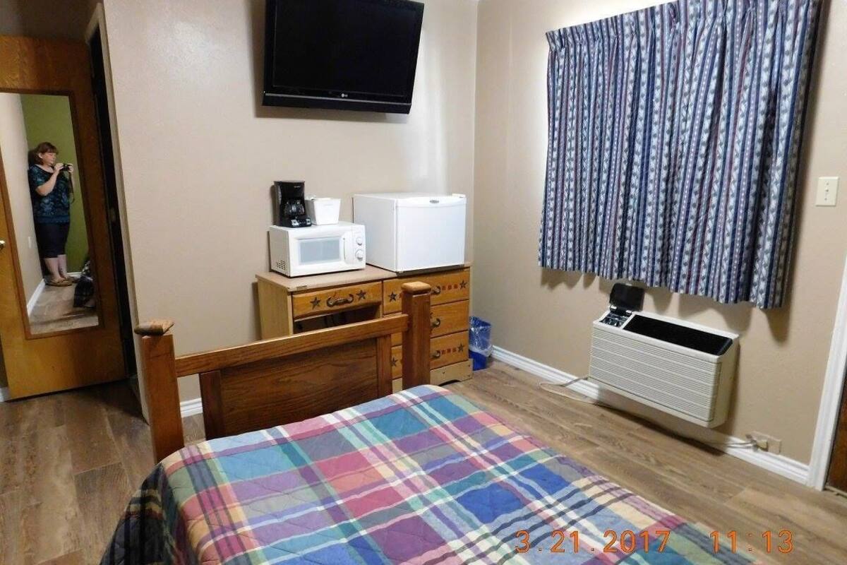 A bedroom with a bed, a dresser, a microwave, a refrigerator, a television, and a window with curtains.