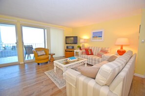 Ein Wohnzimmer mit einer Couch, einem Sessel, einem Couchtisch, einem Fernseher und einem Balkon mit einem Stuhl.