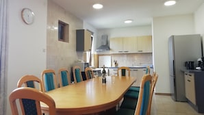 Un comedor con una larga mesa de madera y sillas con cojines azules, una cocina con armarios de colores claros y un reloj en la pared.