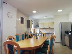 A dining area with a long wooden table and blue cushioned chairs, a kitchen with light-colored cabinets, and a clock on the wall. - Great terrace and panorama view (Nin)