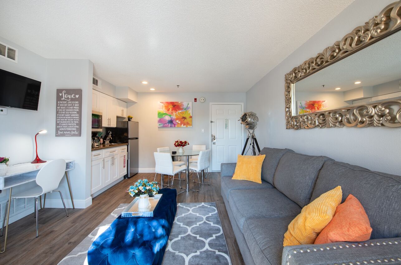 A modern living room with a grey sofa, a blue ottoman, and a dining area with a white table and chairs.