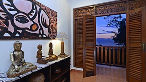 Ein Zimmer mit Buddha-Statue, einem Holzkabinett, einem Gemälde und Blick durch offene Türen nach draußen.