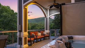 A hot tub area with a wooden deck, outdoor seating, and a view of a forested hillside. - Waterfall Manor | Mountain Luxury w/ Fireplaces & Views + Biltmore Pass (Fletcher)