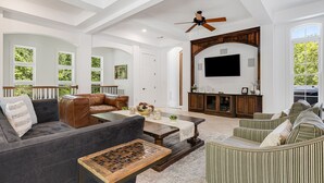 A spacious living room with a ceiling fan, a large flat-screen TV mounted on a wooden cabinet, and a view of greenery through large windows. - Waterfall Manor | Mountain Luxury w/ Fireplaces & Views + Biltmore Pass (Fletcher)