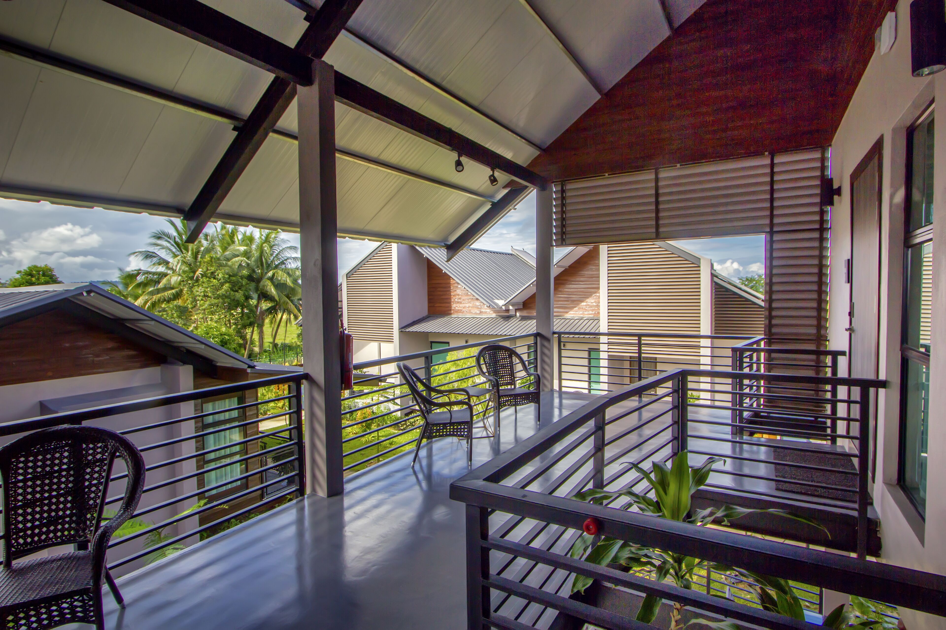 A covered balcony with chairs and a railing, overlooking a residential area with houses and palm trees.