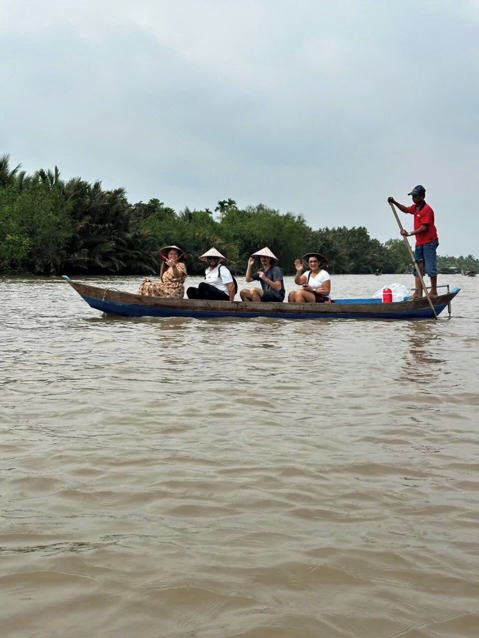 A boat with four people, one person standing at the back, and a calm water body with greenery in the background.