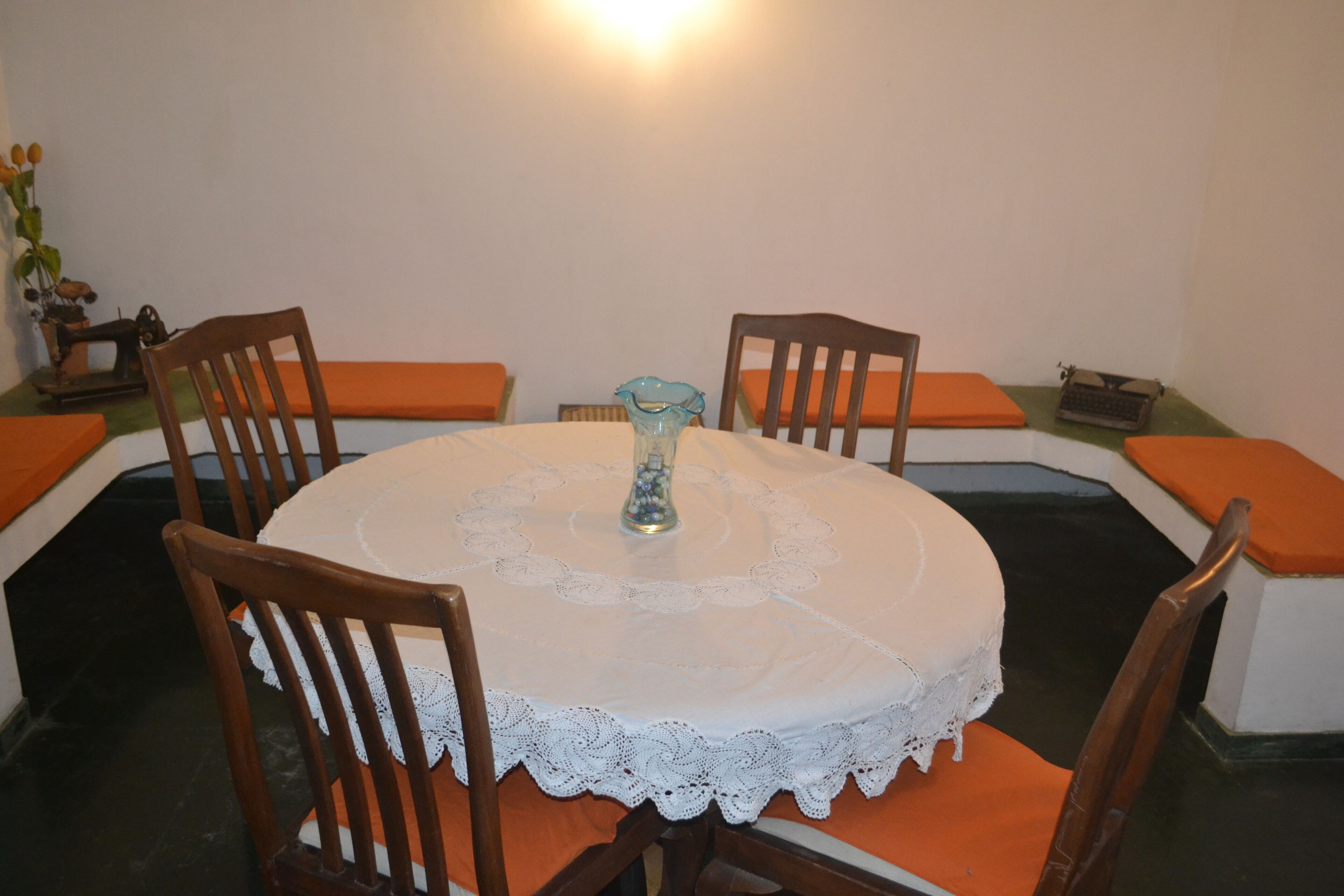 A round dining table with a lace tablecloth, surrounded by wooden chairs with orange cushions.