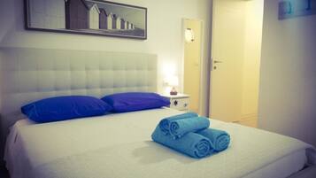 Un letto rifatto con cuscini e asciugamani blu, un comodino con una lampada e un quadro appeso al muro.