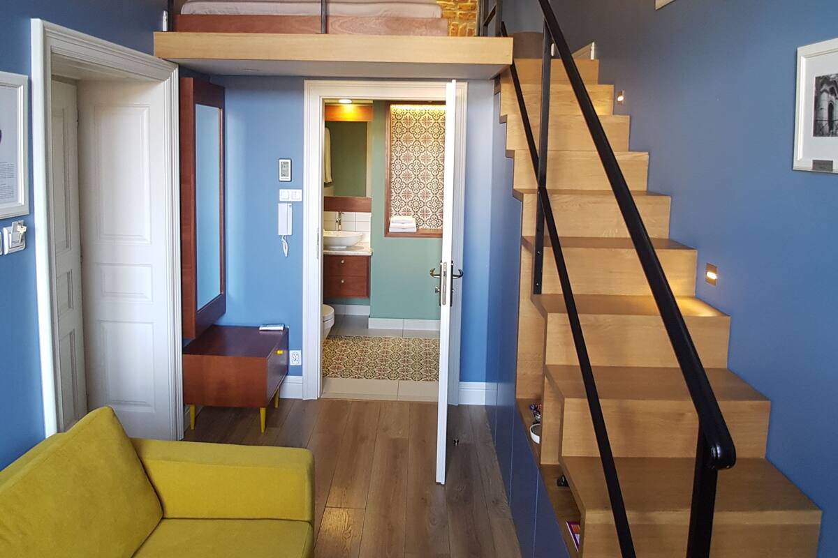 Un intérieur moderne avec un escalier en bois, un canapé jaune et une chambre à l’étage avec un mur en pierre.