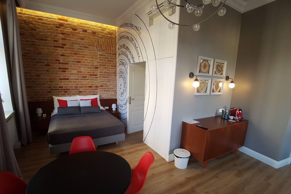 Une chambre moderne avec un mur en briques, un lit avec une literie rouge et blanche, un bureau en bois et une table ronde avec des chaises rouges.