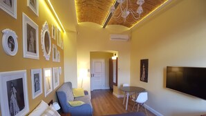 Quarto de hotel moderno com cama, área de estar com mesa e cadeiras, TV na parede e quadros nas paredes.