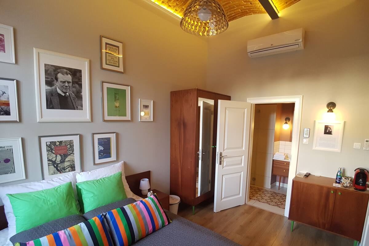 Une chambre à coucher avec un lit, des oreillers verts, un coussin rayé aux couleurs vives, une armoire en bois, un bureau avec une lampe et des tableaux encadrés accrochés au mur.