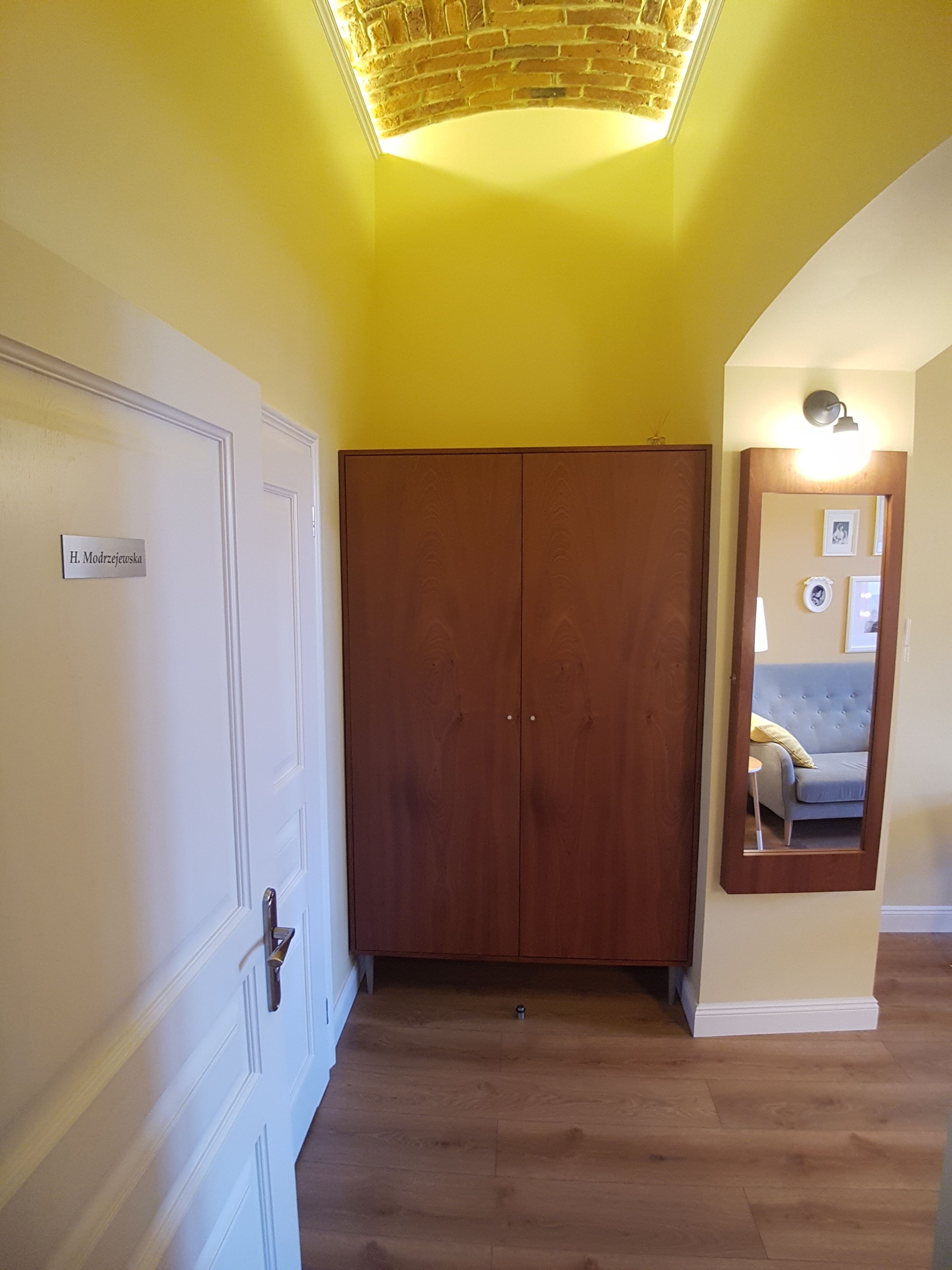 Un pasillo con un armario de madera, una puerta con placa de identificación y un espejo que refleja un sofá y una lámpara.