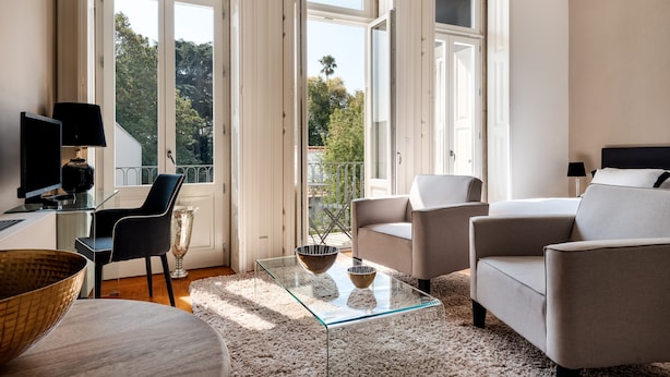 Una sala de estar moderna con una mesa de centro de vidrio, dos sillones, un sofá y un televisor sobre un mueble. Amplias ventanas ofrecen vistas a árboles y un balcón.