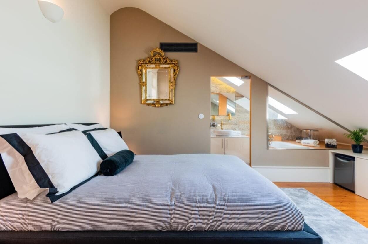 A bedroom with a large bed, a mirror, and a view of a kitchen and bathroom through an angled ceiling.